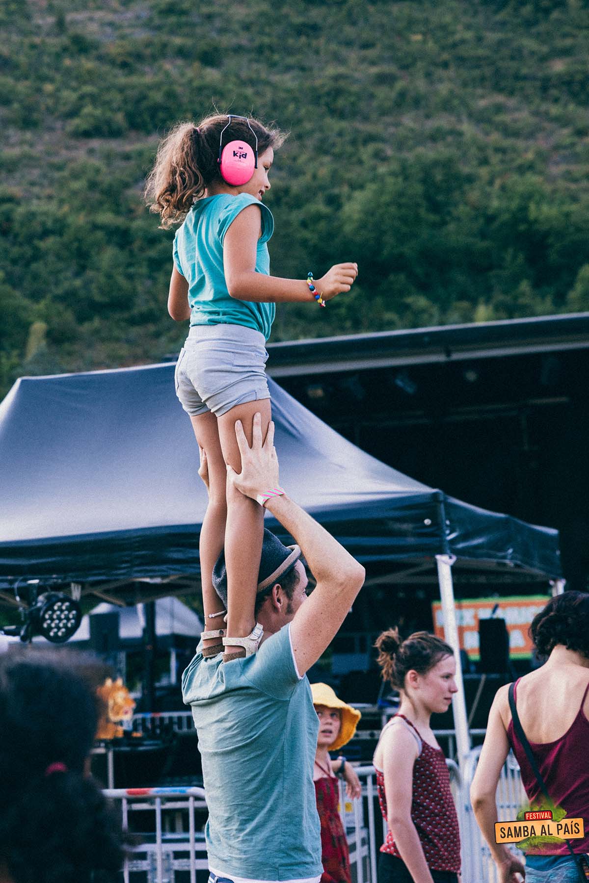 Une petite fille avec un casque anti-bruit rose debout sur les épaules de son père devant la scène d'un concert en fin d'après-midi à Samba al País