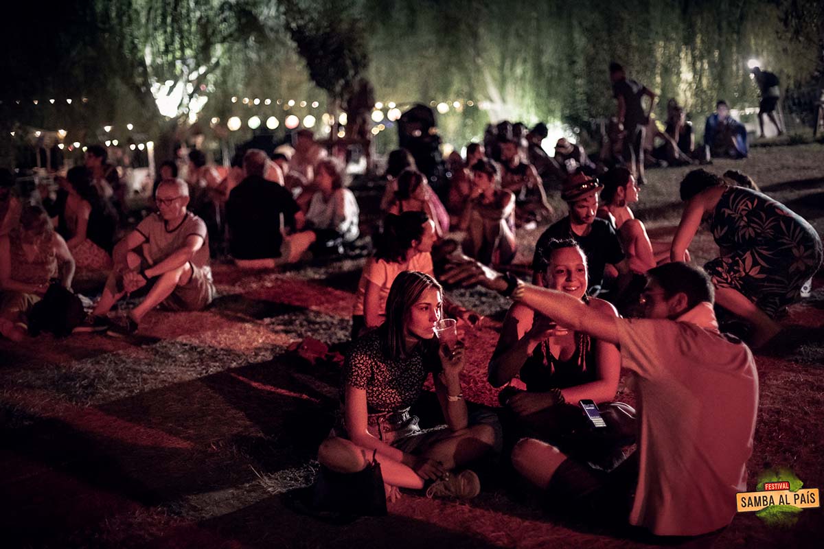 Des festivaliers assis en cercle sur l'herbe qui discutent le soir au festival Samba al País