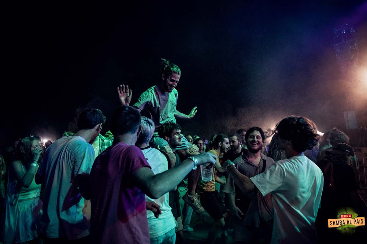 Un jeune homme avec un chignon porté sur les épaules de son ami au milieu d'une foule éclairé par la lumière verte du concert à Samba al País